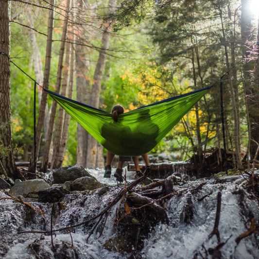 Picture of two people sitting in a hammock in a forest over a stream.